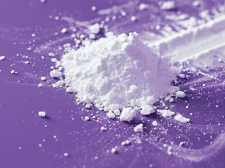 A mound of fine white biotin powder beside a scoop on a purple surface.